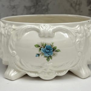 May include: Cream-coloured ceramic planter with a floral design. The planter has a rounded shape, three feet, and ornate detailing. A blue rose is centered on the front. Suitable for indoor use.