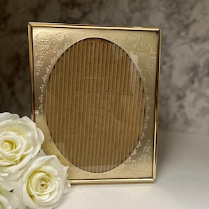May include: A rectangular gold-toned metal picture frame featuring an oval-shaped centre opening with an embossed floral pattern along the corners and edges. The frame contains a piece of brown corrugated cardboard behind the clear glass front.