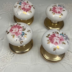May include: A set of four round ceramic cabinet knobs featuring a white base with a painted pink rose floral pattern, blue accents, and gold leaf detailing. Each knob is mounted on a circular brass-toned metal base with a polished finish.