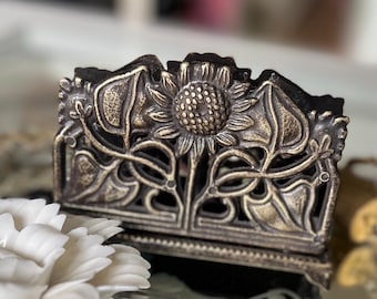 Puede incluir: Un servilletero de metal en tono bronce oscuro con un diseño de girasol en relieve que presenta pétalos y hojas detallados. La base rectangular tiene un borde con cuentas y la estructura calada muestra una flor central prominente.