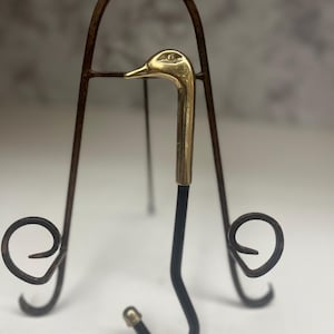 May include: A decorative stand with a brass swan-head handle and a black, curved hook. The stand is made of dark, wrought iron and has a three-legged design with decorative scrollwork.