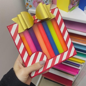 Könnte beinhalten: Eine Geschenkbox mit rot-weißem Streifenmuster, die ein Set bunter Kerzen enthält. Die Box ist mit einer goldenen Schleife verziert. Die Kerzen sind in den Farbtönen Orange, Rosa, Lila, Blau und Grün gehalten.