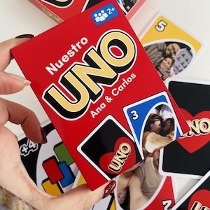 Può includere: Una scatola rossa del gioco di carte UNO con la scritta "Nuestro UNO Ana & Carlos" e un mazzo di carte. Le carte hanno foto di persone. Il gioco è per 2+ giocatori.