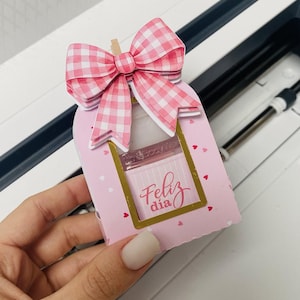 Puede incluir: Una caja de regalo rosa con un lazo de cuadros y las palabras "Feliz dia" en cursiva. La caja tiene una ventana con marco dorado y pequeños detalles de corazón. La caja es sostenida por una persona.