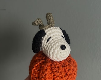 Op de afbeelding: Een handgemaakt gehaakt Snoopy-personage bovenop een oranje pompoen. Het Snoopy-figuur heeft een witte kop, zwarte oren en een zwarte neus. De pompoen is fel oranje van kleur en het Snoopy-figuur heeft een kleine, beige stengel.