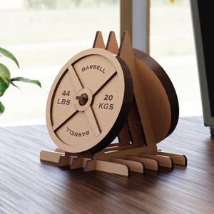 Może przedstawiać: Drewniany design talerza do sztangi z napisem "BARBELL" i pomiarem wagi 20 kg i 44 lbs. Dekoracyjny przedmiot jest wykonany z jasnobrązowego drewna i stoi na biurku.