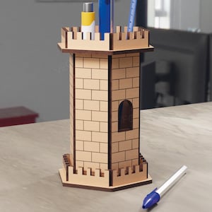 May include: A wooden pen holder shaped like a castle tower. The tower is light brown with a brick pattern and a small arched window. It holds pens, a ruler, and a small bottle. A pen with a blue tip rests on the table.