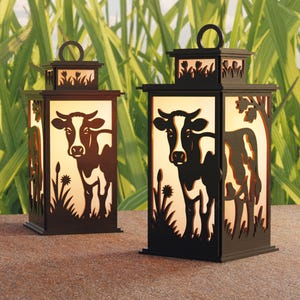 Può includere: Due lanterne decorative in metallo nero con silhouette di mucche. Ogni lanterna ha una forma rettangolare con un manico in alto e disegni ritagliati di mucche, erba e fiori. Le lanterne sono illuminate dall'interno.