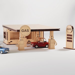 Op de afbeelding: Een houten model van een benzinestation met een rode auto onder de luifel. Een blauwe auto staat in de buurt geparkeerd. Het benzinestation heeft een bord met de tekst "GAS". Het model is gemaakt van lichtgekleurd hout.