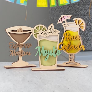 May include: Wooden cocktail decorations, including an Espresso Martini, Mojito, and Pi&ntilde;a Colada. Each drink is laser-cut with detailed designs and text, ideal for a bar or party. The Mojito is green, the Pi&ntilde;a Colada is yellow, and the Espresso Martini is brown.