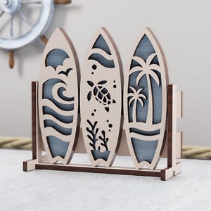 Puede incluir: Decoración de tabla de surf de madera con tres tablas de surf en un soporte. Cada tabla de surf presenta un diseño recortado único: olas, una tortuga y palmeras. El soporte es marrón y las tablas de surf son de color beige claro.