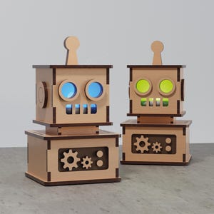 Può includere: Due lampade in legno a forma di robot con occhi e bocche illuminate. I robot hanno una finitura marrone chiaro con ritagli a forma di ingranaggio sulla base. Un robot ha gli occhi blu, l'altro gli occhi verdi.