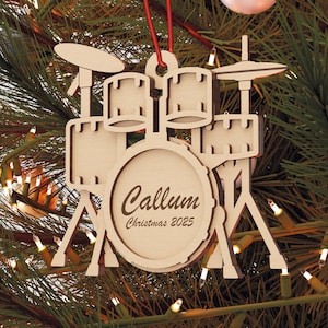 Puede incluir: Adorno navideño de batería de madera con el nombre "Callum" y el año "Christmas 2025" grabado en el bombo. El adorno cuelga de un árbol de Navidad con luces y otros adornos.