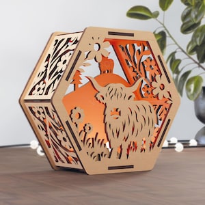 Könnte beinhalten: Eine sechseckige Holzlaterne mit einer Highland-Kuh-Silhouette und floralen Ausschnitten. Das Innere leuchtet mit orangefarbenem Licht und erzeugt eine warme Atmosphäre. Die Laterne steht auf einer Holzoberfläche.
