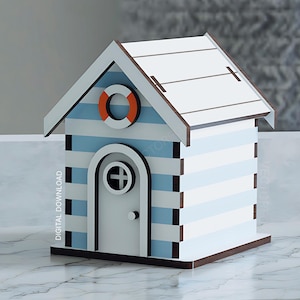 Op de afbeelding: Een decoratief houten strandhuisje met blauw-wit gestreepte muren, een wit dak en een kleine deur. Het heeft een reddingsboei en is een charmant nautisch item.
