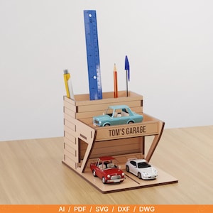 Puede incluir: Un organizador de escritorio de madera con forma de garaje, con el texto "TOM'S GARAGE". Contiene bolígrafos, lápices y una regla azul. Incluye coches de juguete: un coche clásico rojo, un coche deportivo blanco y un coche vintage azul claro.