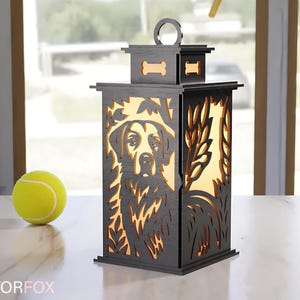 May include: A wooden lantern with a dog silhouette design. The lantern is dark gray with a light-colored interior, featuring a dog's face and bone-shaped cutouts. A yellow tennis ball sits nearby.