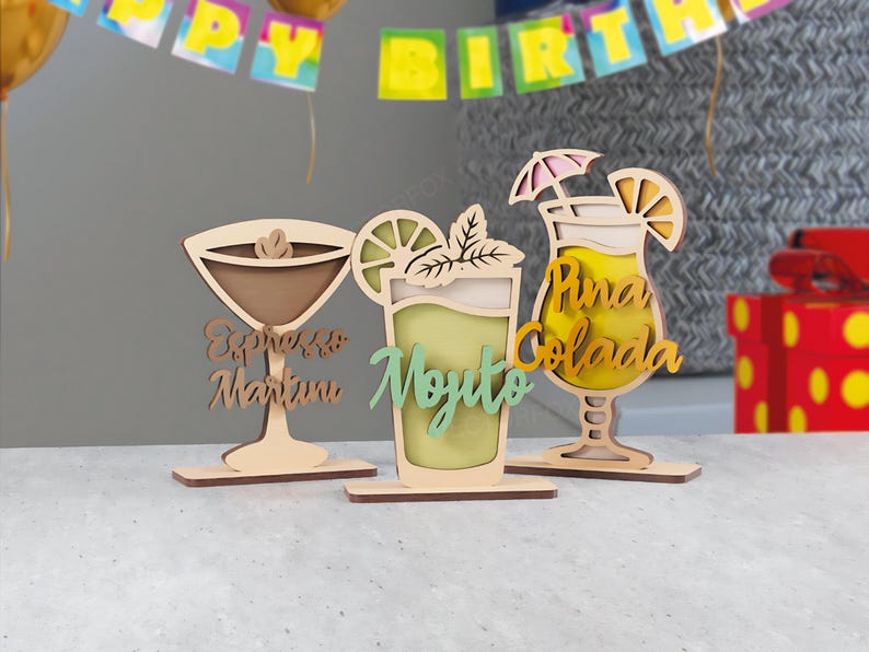 May include: Wooden cocktail decorations, including an Espresso Martini, Mojito, and Pi&ntilde;a Colada. Each drink is laser-cut with detailed designs and text, ideal for a bar or party. The Mojito is green, the Pi&ntilde;a Colada is yellow, and the Espresso Martini is brown.