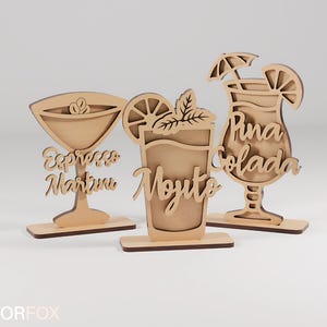 May include: Three wooden cocktail decorations: an Espresso Martini, a Mojito, and a Pi&ntilde;a Colada. Each drink is laser-cut from light-coloured wood and features the drink's name in cursive script. The Mojito has a mint sprig and a lemon slice.