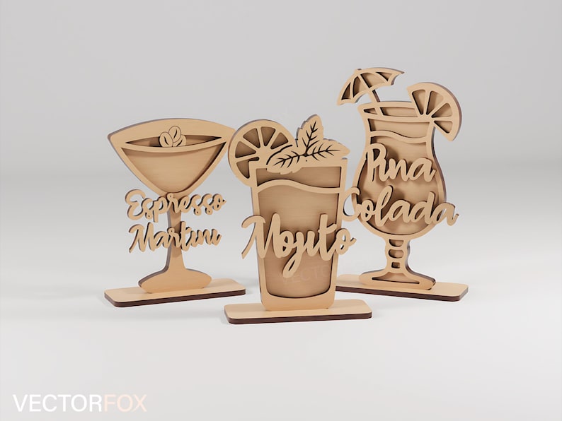 May include: Three wooden cocktail decorations: an Espresso Martini, a Mojito, and a Pi&ntilde;a Colada. Each drink is laser-cut from light-coloured wood and features the drink's name in cursive script. The Mojito has a mint sprig and a lemon slice.