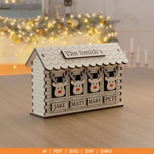 Op de afbeelding: Een lichtkleurig houten ornament in de vorm van een schuur met vier rendierkoppen in stallen. Het dak is gelabeld met The Smith's en de vier stallen zijn gepersonaliseerd met de namen JAKE, MATT, MARY en PETE in zwarte letters op de voorpanelen.