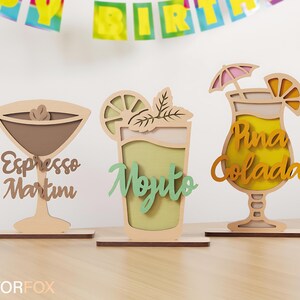 May include: Wooden cocktail decorations, including an espresso martini, mojito, and pi&ntilde;a colada. Each drink is laser-cut with layered details and the drink's name. The decorations are on wooden bases.