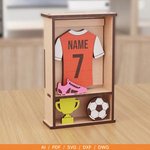 Könnte beinhalten: Eine hölzerne Vitrine mit einem Ausschnitt eines orange-weißen Fußballtrikots mit dem Text NAME und der Nummer 7, einem rosa Fußballschuh, einem gelben Pokal und einem schwarz-weißen Fußball. Am unteren Rand steht AI, PDF, SVG, DXF, DWG.