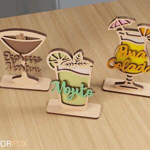 May include: Three wooden cocktail-themed decorations. The first is a martini glass with the text "Espresso Martini". The second is a mojito with the text "Mojito". The third is a pi&ntilde;a colada with the text "Pina Colada".