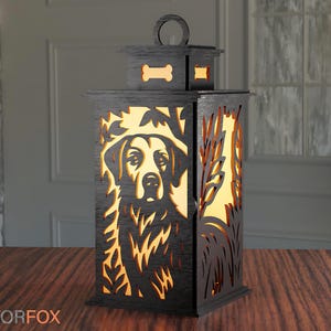 May include: A black wooden lantern with a dog portrait cutout design. The lantern features a handle, a bone-shaped cutout, and a rectangular shape. The dog's face is visible through the cutout, and the lantern is placed on a wooden surface.