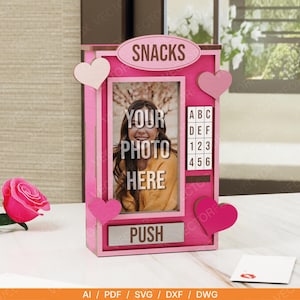 May include: A pink wooden snack machine-themed photo frame with the word "SNACKS" at the top. The frame has a photo of a person with the text "YOUR PHOTO HERE". It also has a keypad and the word "PUSH". Decorative pink hearts are attached to the frame.