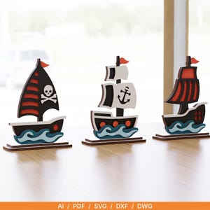 May include: Three decorative wooden ship cutouts featuring a pirate vessel with a skull and crossbones, a ship with an anchor emblem, and a vessel with red sails. Each ship is mounted on a base and displays stylized blue waves at the hull.