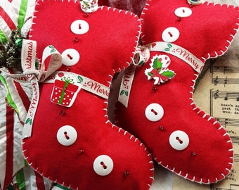 May include: Red felt Christmas stocking ornaments with white stitching, white buttons, and decorative ribbons that say "Merry". One stocking has a gift box embellishment, and another has a holly and candy cane embellishment. Festive holiday decor.