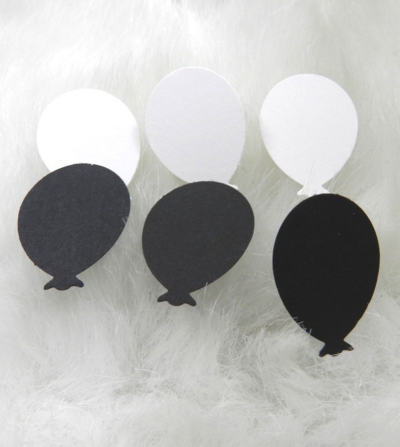 May include: Three pairs of black and white balloon shaped paper cutouts. The balloons are arranged in a row on a white fluffy surface.