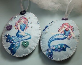 May include: Two blue and white felt ornaments with a mermaid design. Each ornament has a white ribbon for hanging and is decorated with pearl-like beads and colorful buttons.