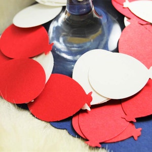 May include: Red and white paper balloon cutouts arranged around a clear glass stemware. The balloons are layered and overlapping, creating a festive and celebratory look.