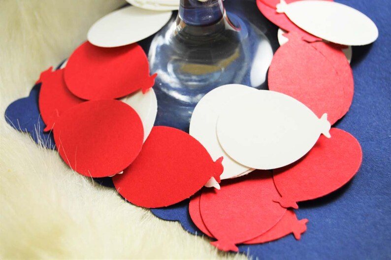 May include: Red and white paper balloon cutouts arranged around a clear glass stemware. The balloons are layered and overlapping, creating a festive and celebratory look.