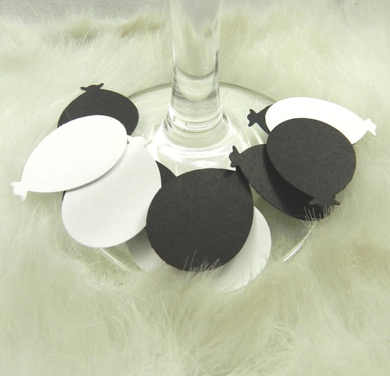 May include: Black and white paper balloon confetti scattered around a wine glass. The confetti is shaped like balloons with a rounded bottom and a pointed top.