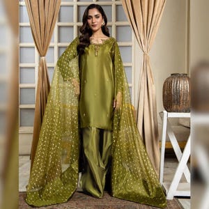 May include: Olive green formal wear ensemble. The outfit includes a long-sleeved tunic with subtle embellishments, matching wide-leg trousers, and a sheer, flowing cape adorned with gold polka dots. The fabric has a satin sheen.