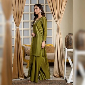 May include: Olive green satin two-piece outfit. The long-sleeved tunic features delicate embroidery and a matching wide-leg pant. The outfit is worn with a matching scarf. The background includes gold curtains and a decorative vase.