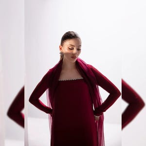 May include: A burgundy long-sleeved dress with a sheer, matching shawl. The dress features a decorative, embellished neckline. The outfit is a formal style, suitable for special occasions. The model is wearing earrings.