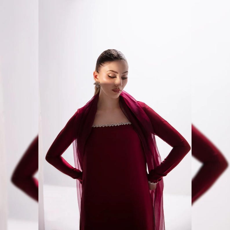May include: A burgundy long-sleeved dress with a sheer, matching shawl. The dress features a decorative, embellished neckline. The outfit is a formal style, suitable for special occasions. The model is wearing earrings.