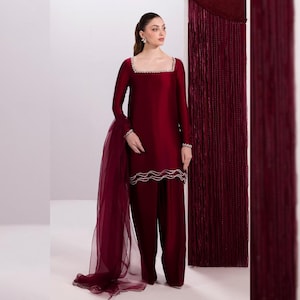 May include: A burgundy two-piece outfit featuring a long-sleeved top with a square neckline and embellished trim, paired with matching wide-leg pants. A sheer scarf drapes from the shoulder. The outfit is a solid color, with a flowing silhouette.