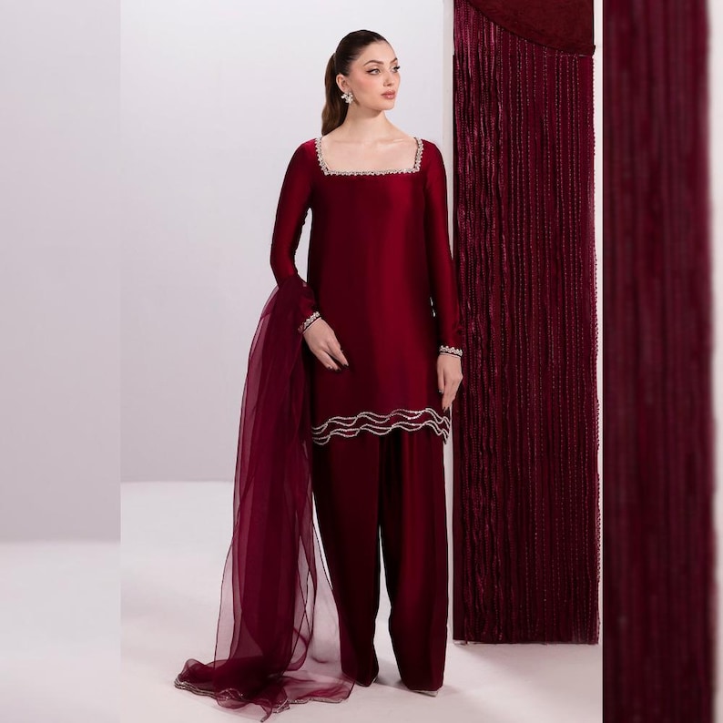 May include: A burgundy two-piece outfit featuring a long-sleeved top with a square neckline and embellished trim, paired with matching wide-leg pants. A sheer scarf drapes from the shoulder. The outfit is a solid color, with a flowing silhouette.