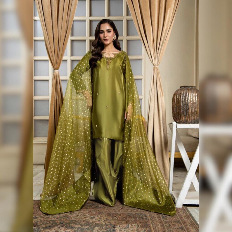 May include: Olive green formal wear ensemble. The outfit includes a long-sleeved tunic with subtle embellishments, matching wide-leg trousers, and a sheer, flowing cape adorned with gold polka dots. The fabric has a satin sheen.