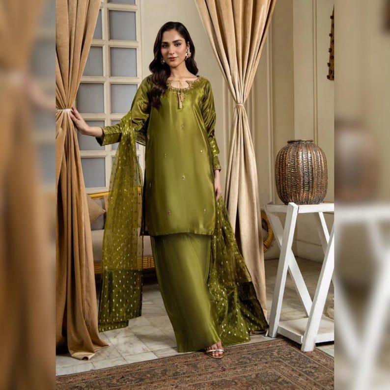 May include: Olive green satin salwar kameez ensemble. The outfit includes a long-sleeved tunic with gold embellishments, matching trousers, and a sheer dupatta with gold polka dots. The woman is standing in front of a window with beige curtains.