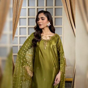 May include: An olive green satin dress with long sleeves and a sheer overlay with gold polka dots. The dress has a keyhole neckline with gold embroidery and small gold embellishments. The model is wearing a floral earring.