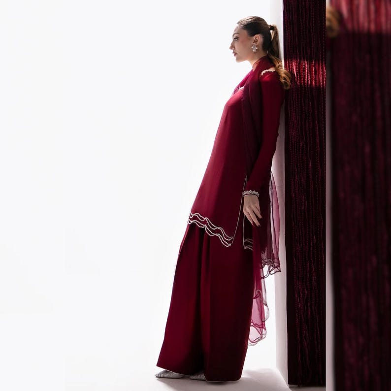 May include: A burgundy ensemble featuring a long-sleeved tunic and wide-leg pants. The tunic has white wavy trim along the hem and sleeves. A sheer burgundy scarf is draped over the shoulders. The outfit is worn with silver shoes.