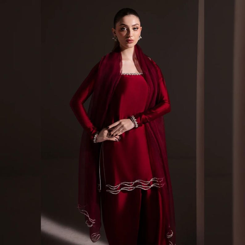 May include: A deep red formal outfit, featuring a long-sleeved top and matching pants. The top has a square neckline and is embellished with a silver trim. A sheer, burgundy shawl drapes over the shoulders. The outfit is accessorized with matching bracelets.