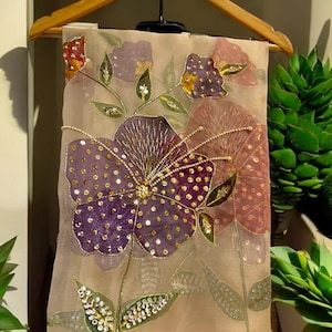 May include: A sheer, beige fabric featuring embroidered floral designs in purple, red, and pink. The flowers are adorned with gold sequins and beads. The fabric is suspended from a wooden hanger.