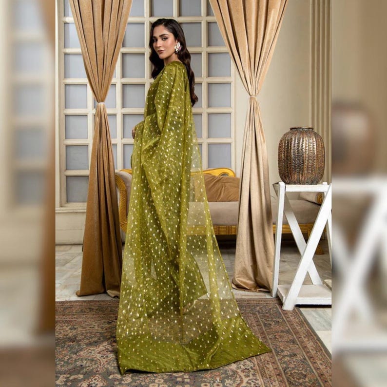 May include: A flowing, olive green dress with a sheer overlay dotted with gold accents. The dress features long sleeves and a draped design, creating an elegant silhouette. The fabric appears lightweight and airy, suitable for formal occasions.
